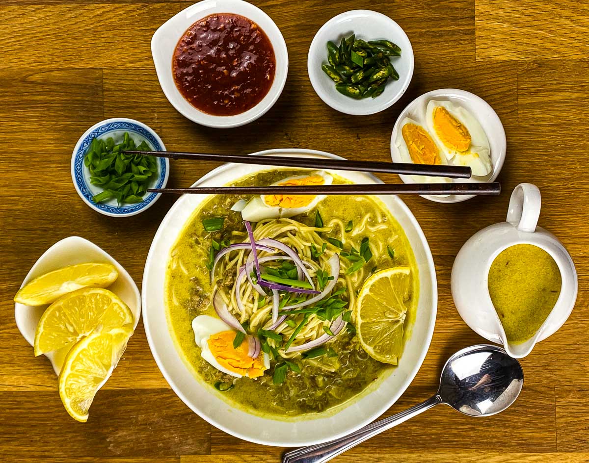 Noodle soup in a bowl with lemon wedges, boiled eggs, cilantro, green chillies, and extra broth