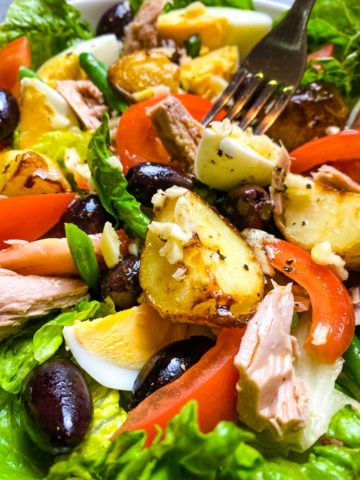 Salad with tuna, olives, dressing, potatoes, lettuce, and tomatoes.