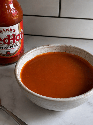 Homemade buffalo sauce in a bowl with Frank’s RedHot sauce and a spoon on a marble countertop.