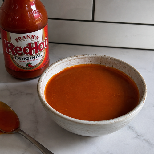 Homemade buffalo sauce in a bowl with Frank’s RedHot sauce and a spoon on a marble countertop.